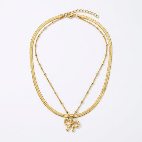 A gold double-strand necklace featuring a textured snake chain and a delicate beaded chain with a small bow pendant, displayed on a clean white background.