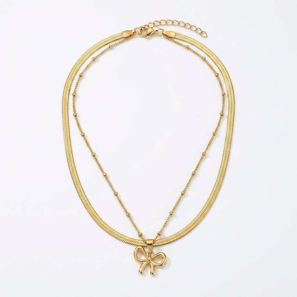 A gold double-strand necklace featuring a textured snake chain and a delicate beaded chain with a small bow pendant, displayed on a clean white background.